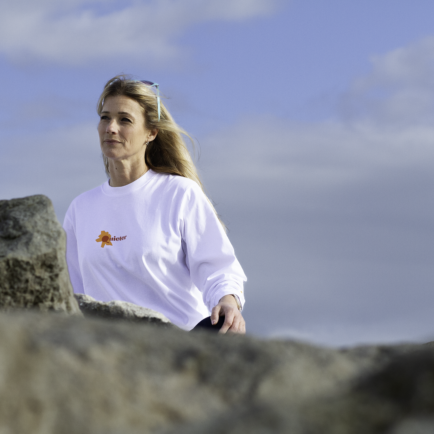Woman in a white shirt standing on a rocky beach with a cloudy sky.