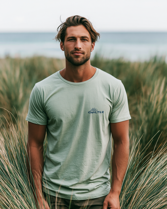 A man wearing a sage green Quieter t-shirt stands in a field of tall beach grass with the ocean in the background.