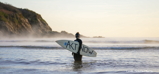 Woman in Ocean holding a surfboard with ACT NOW written on board.
