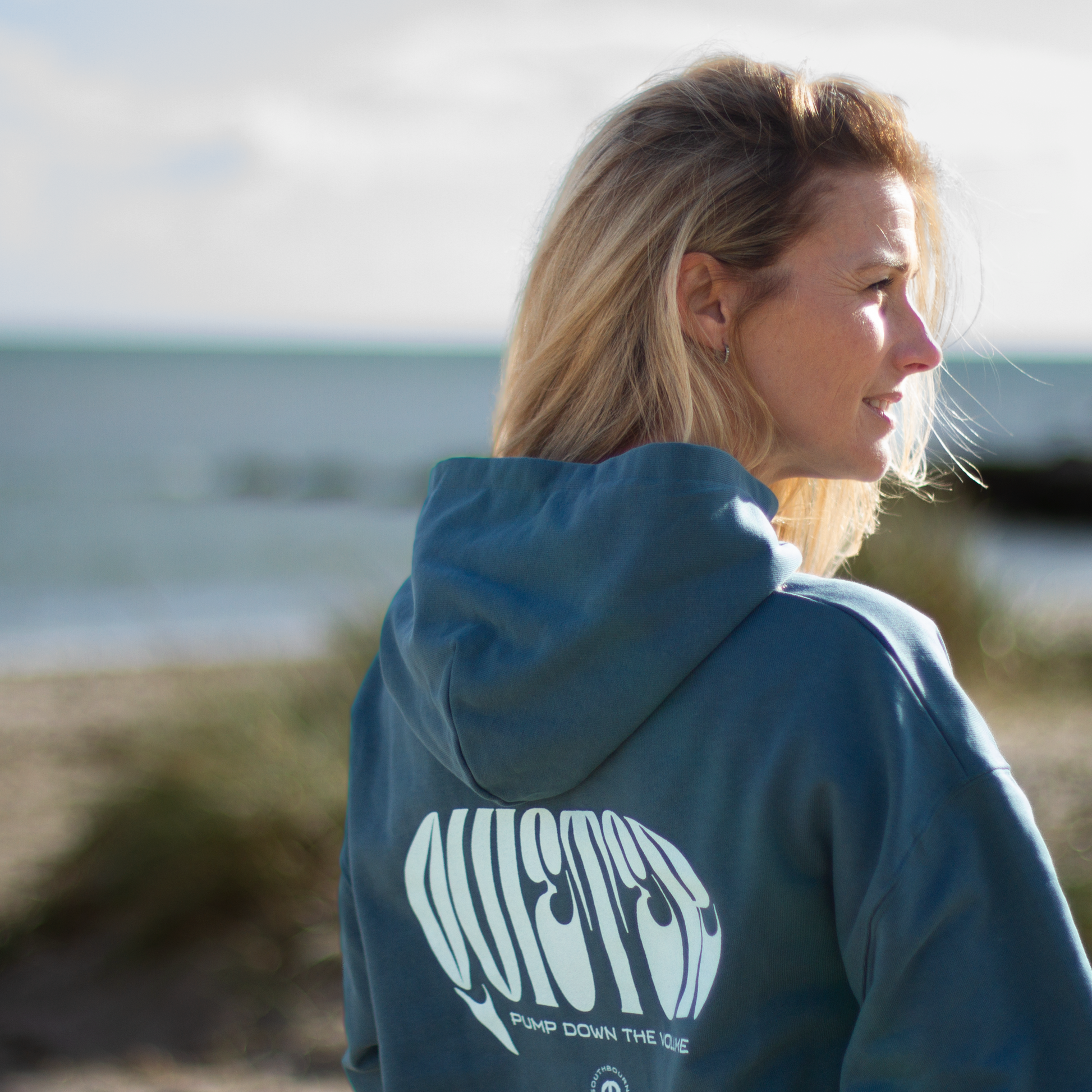 Person wearing a blue hoodie with a logo, standing by a body of water.