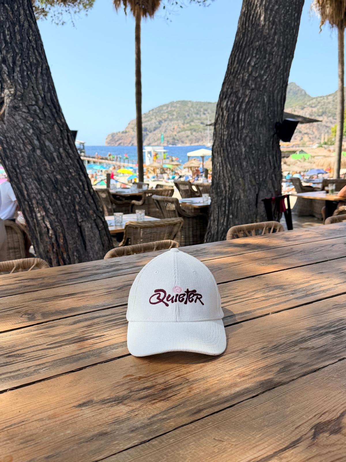 A white baseball cap with 'Quieter' embroidered in black and pink on the front, resting on a rustic wooden table at an outdoor beachside cafe with trees and a beach in the background.