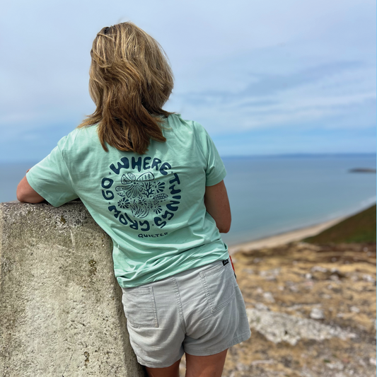A woman viewed from behind wearing a mint green t-shirt with a navy blue circular graphic that reads 'WHERE TWINDS CROW GO' and 'CROWTWINS'. She is leaning on a concrete wall overlooking a coastal landscape with the ocean and beach in the background.