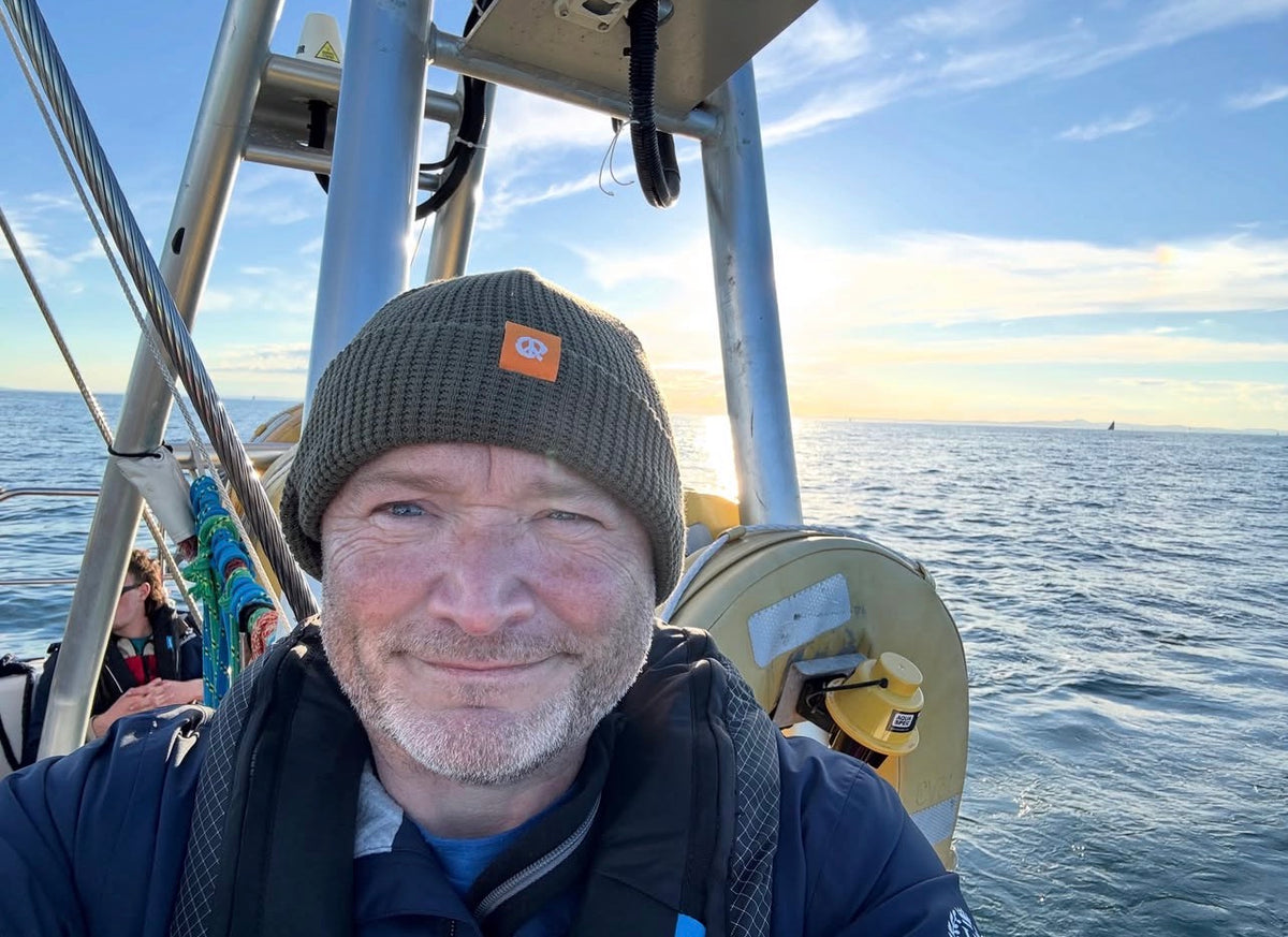 A man wearing a dark olive green knit beanie with an orange logo patch and a blue jacket takes a selfie on a boat with the ocean and sky in the background.