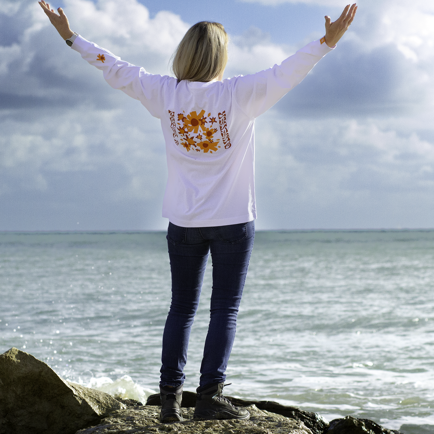 Person standing on a rocky beach with arms outstretched, wearing a white shirt with a floral design.