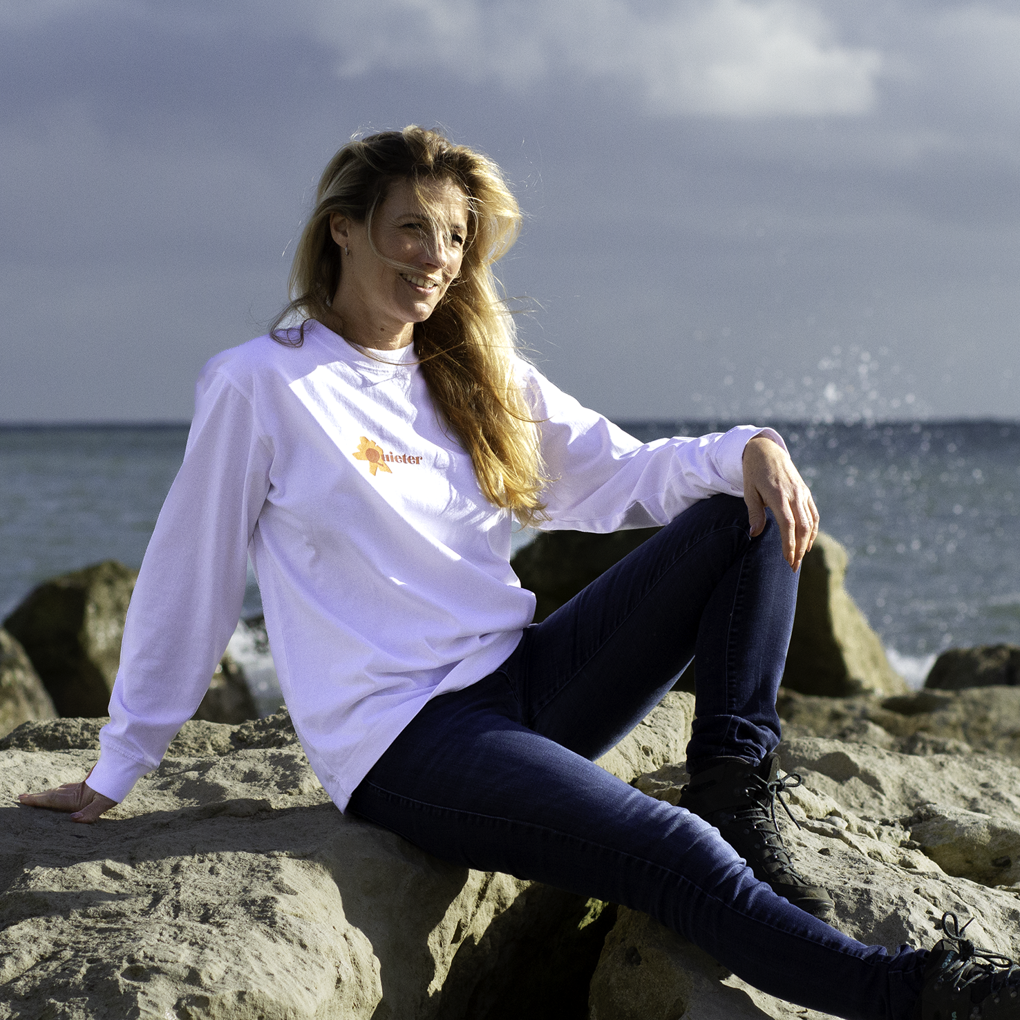 Woman sitting on rocks by the ocean wearing a white shirt with a logo.