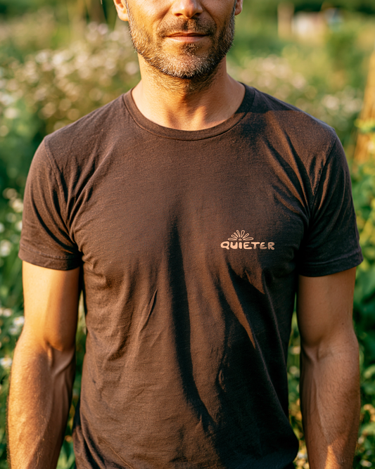 A man wearing a dark brown crew-neck t-shirt with a small 'QUIETER' logo printed on the left chest, set against a blurred green outdoor background.