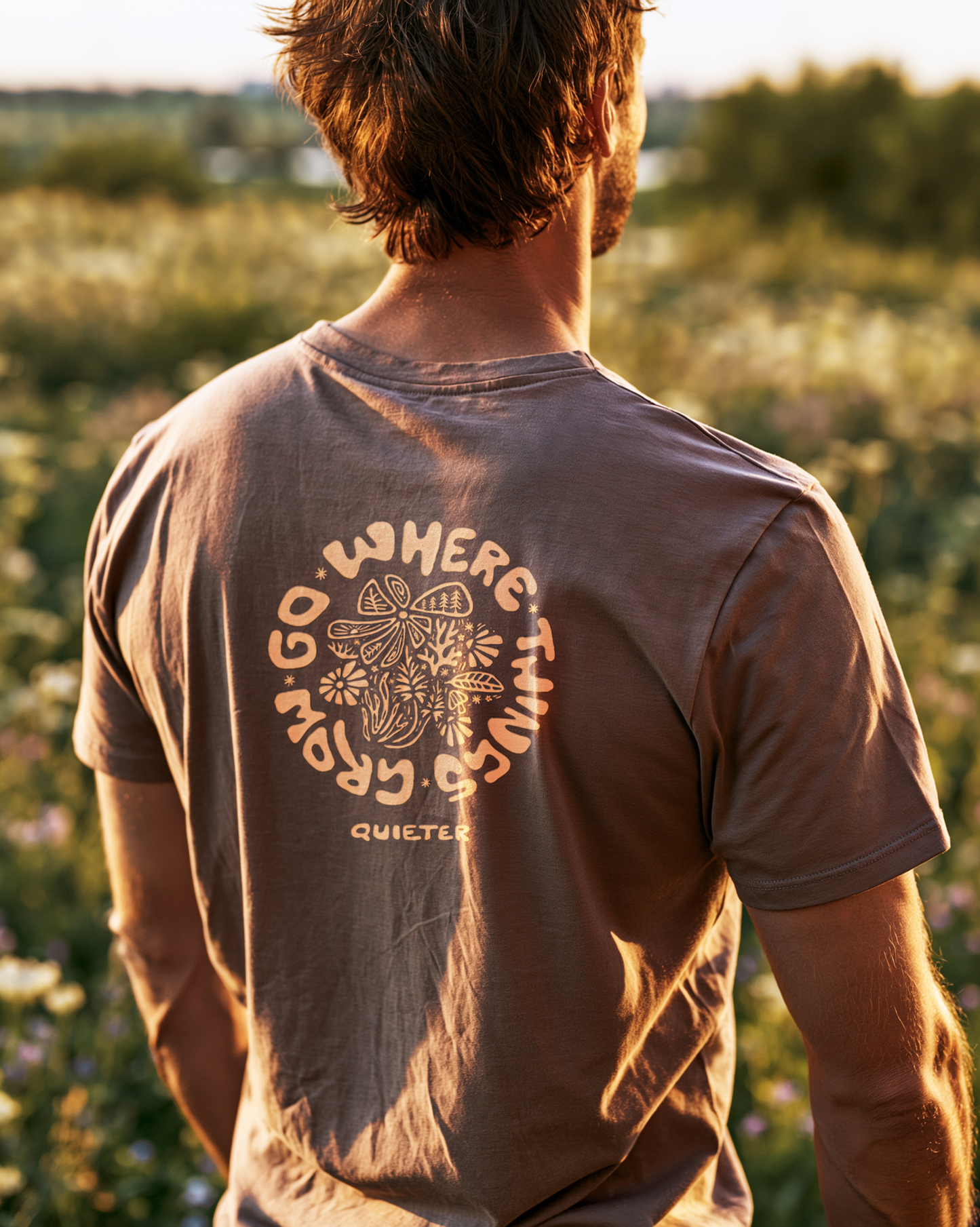 A man viewed from behind wearing a brown t-shirt with a circular graphic on the back that reads 'GO WHERE THINGS GROW' around a mushroom and flower design, with 'QUIETLY' below. The background is a blurred outdoor field at sunset.