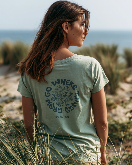 A woman with long brown hair is shown from behind wearing a sage green t-shirt with a dark graphic print on the back. The print features a mandala design with the text 'GO WHERE THINGS GROW' and the brand name 'QUIETER' below it.